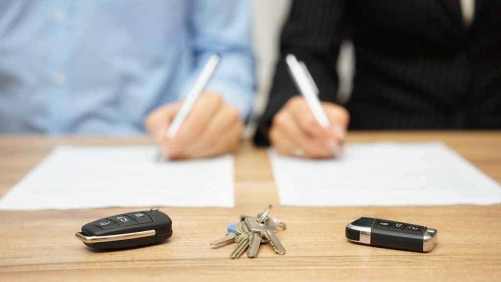 Two people sign separate documents, with car keys and house keys on the table between them