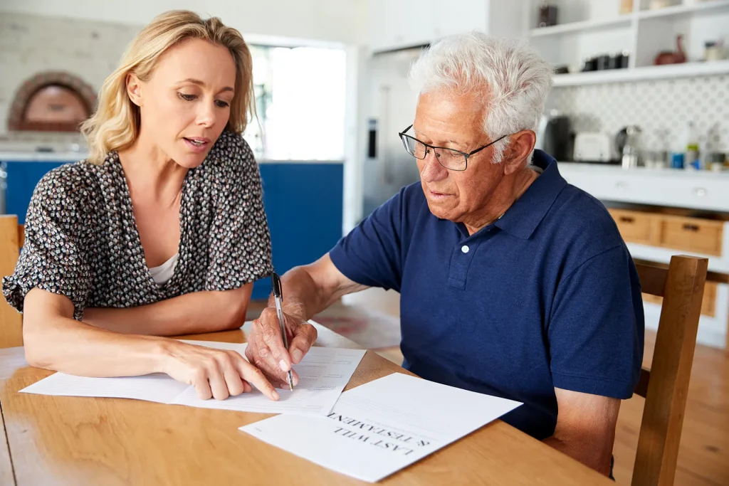 An older man signs a last will and testament at a kitchen table while a younger woman points to the page