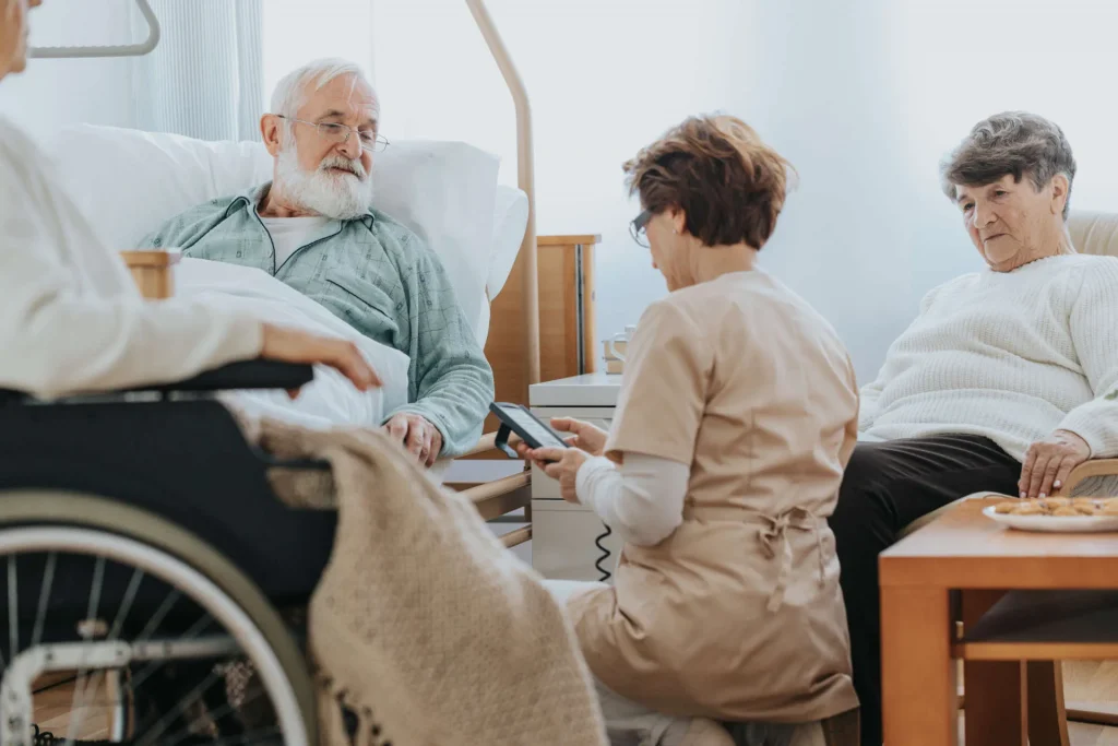 A carer attends an elderly man in a care home, a wheelchair in the foreground
